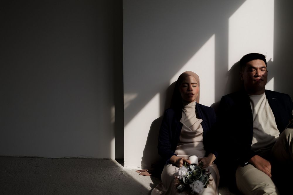 Couple in formal attire sitting on a sofa with soft natural light and shadows, bride holding white flowers.