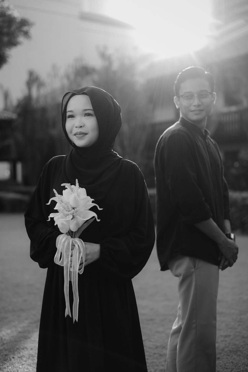 Black and white portrait of Muslim couple, woman in hijab holding white flowers with backlit sunset glow.