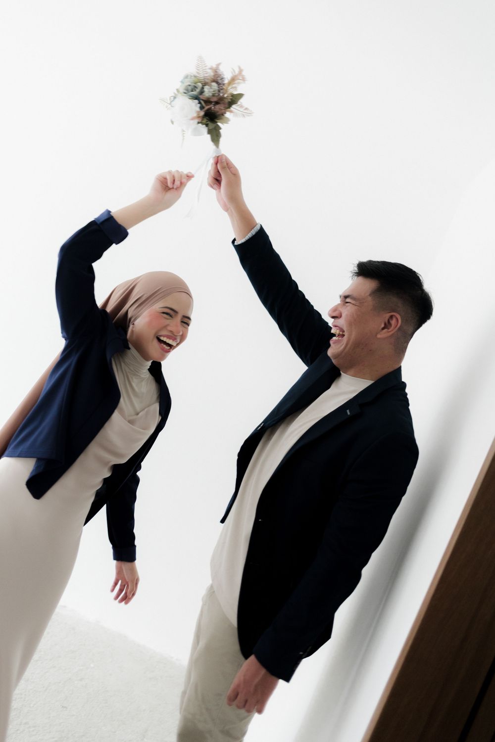 Joyful couple in matching navy blazers laughing while holding a wedding bouquet together against white background.
