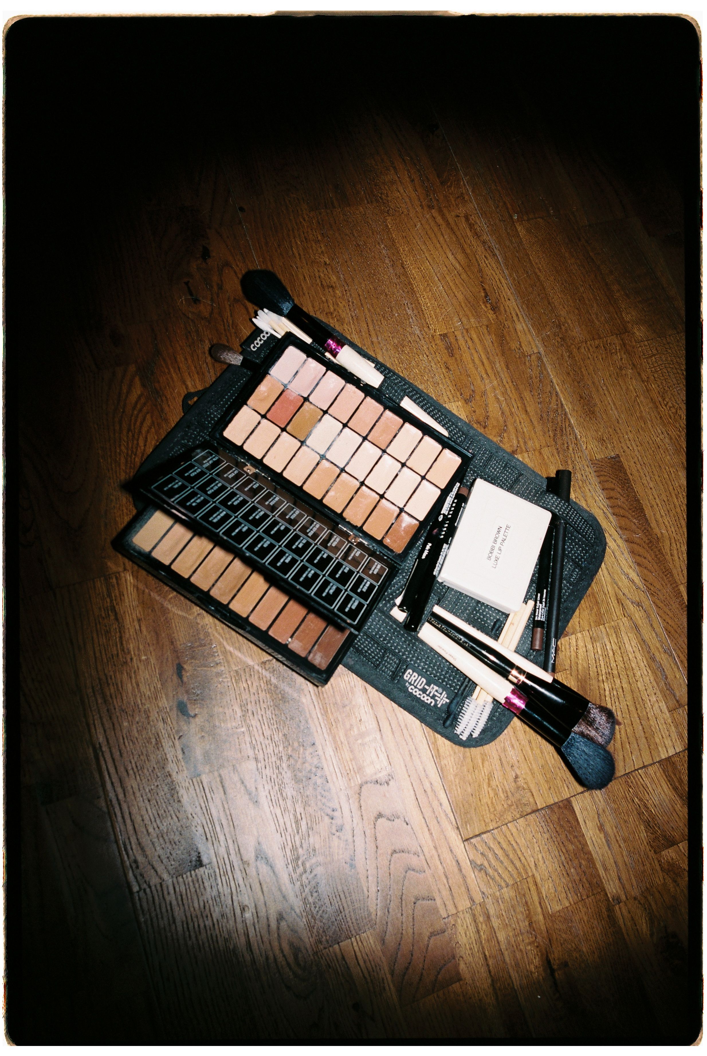 Large makeup palette with neutral eyeshadows, brushes, and beauty tools arranged on a wooden floor.