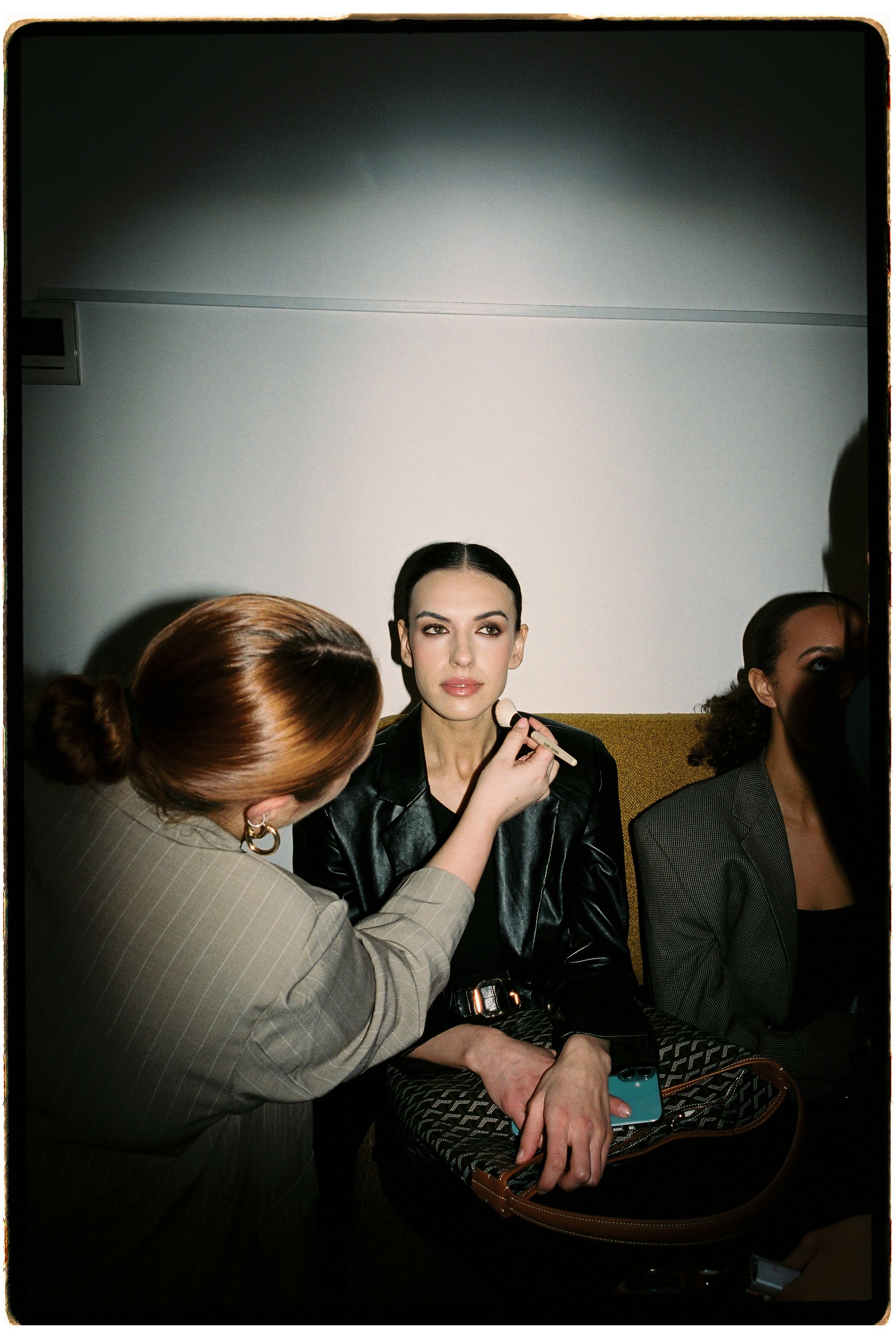 A makeup artist applies finishing touches to a dark-haired model in a black leather jacket backstage at a fashion event.