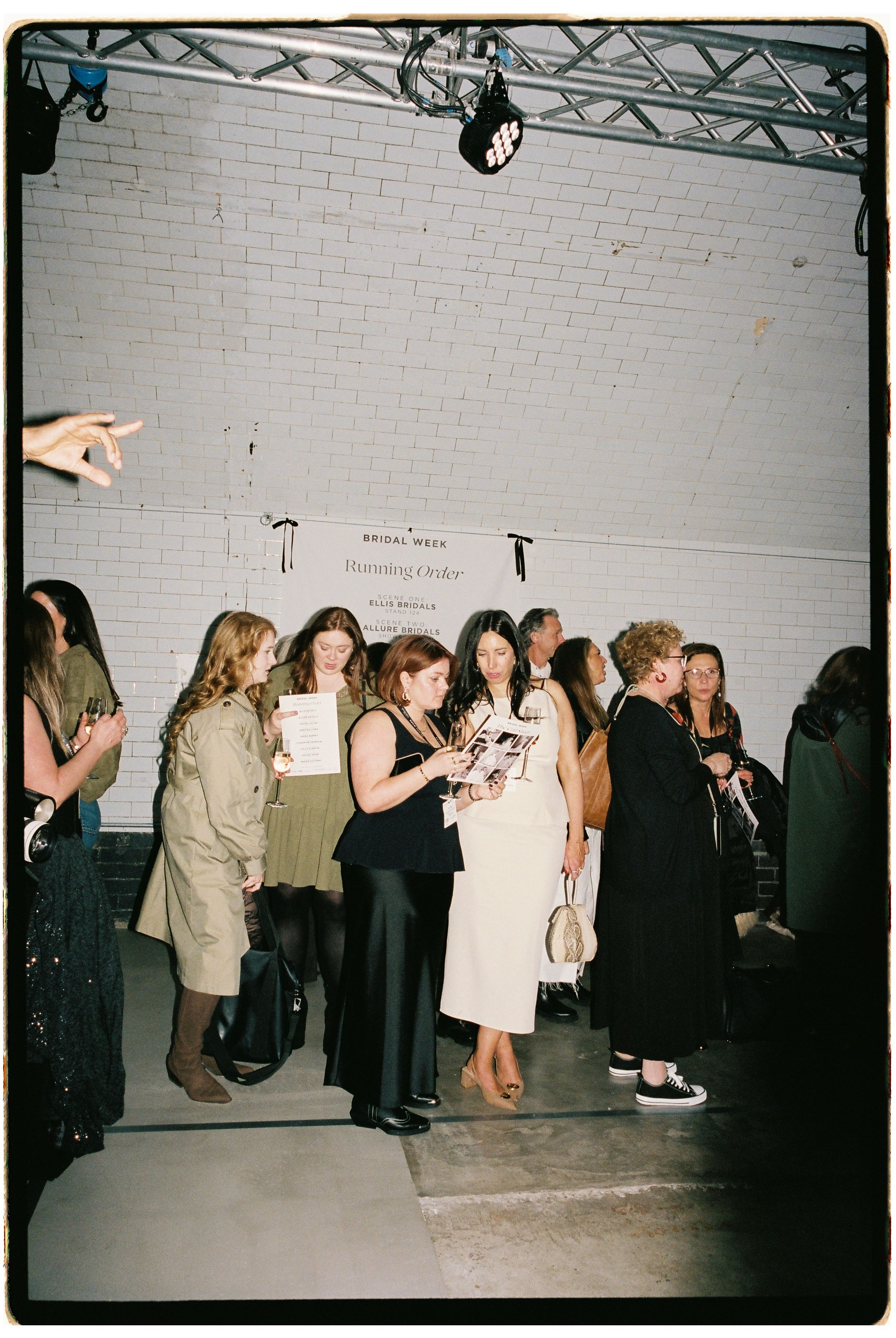 Guests gather at a bridal press event in a white brick venue, browsing magazines and mingling under industrial lighting.