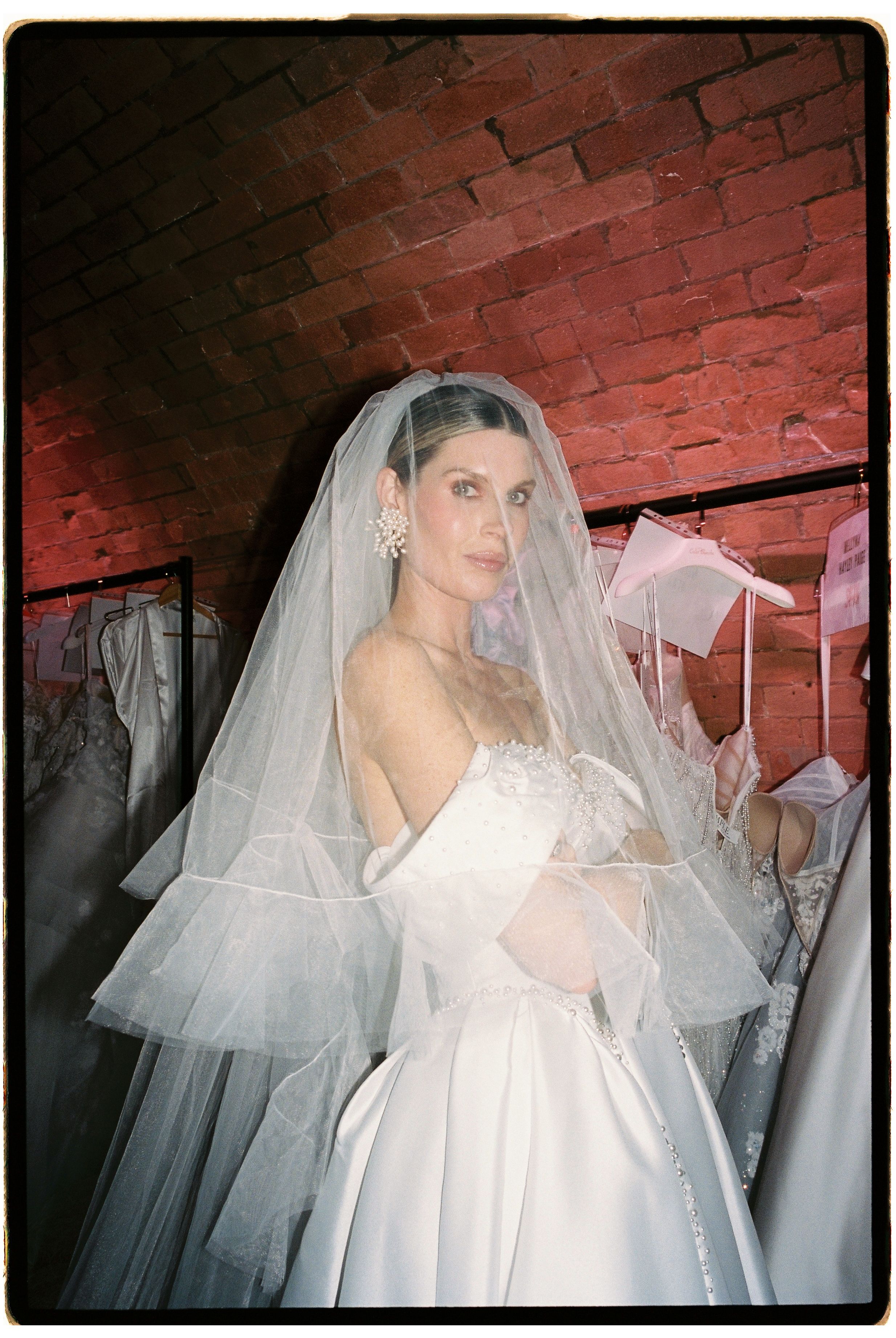 Bride in elegant white ball gown and layered veil poses in front of red brick wall surrounded by wedding dresses.