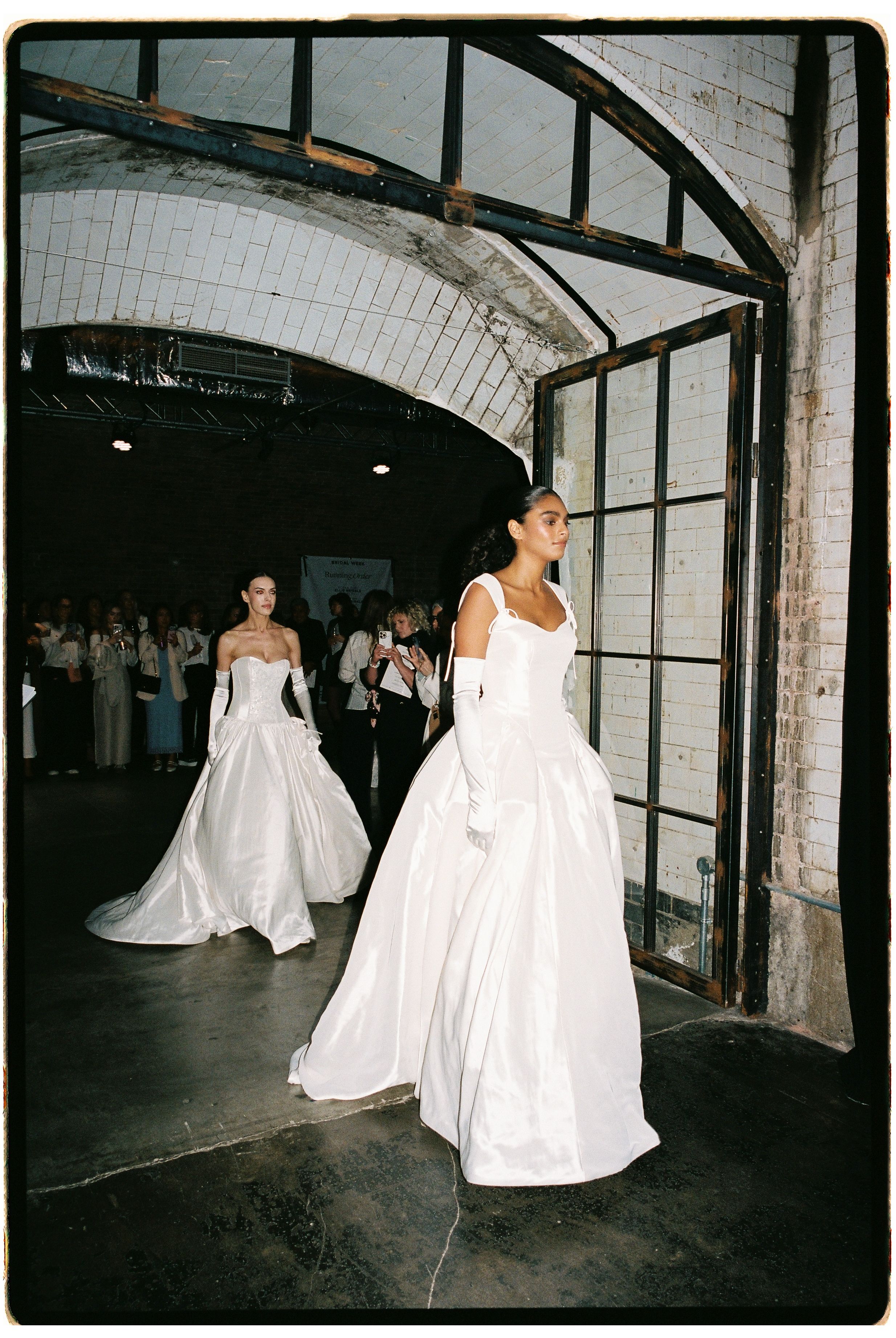 Two models walk in white ball gowns with gloves at a bridal fashion show in an industrial venue.
