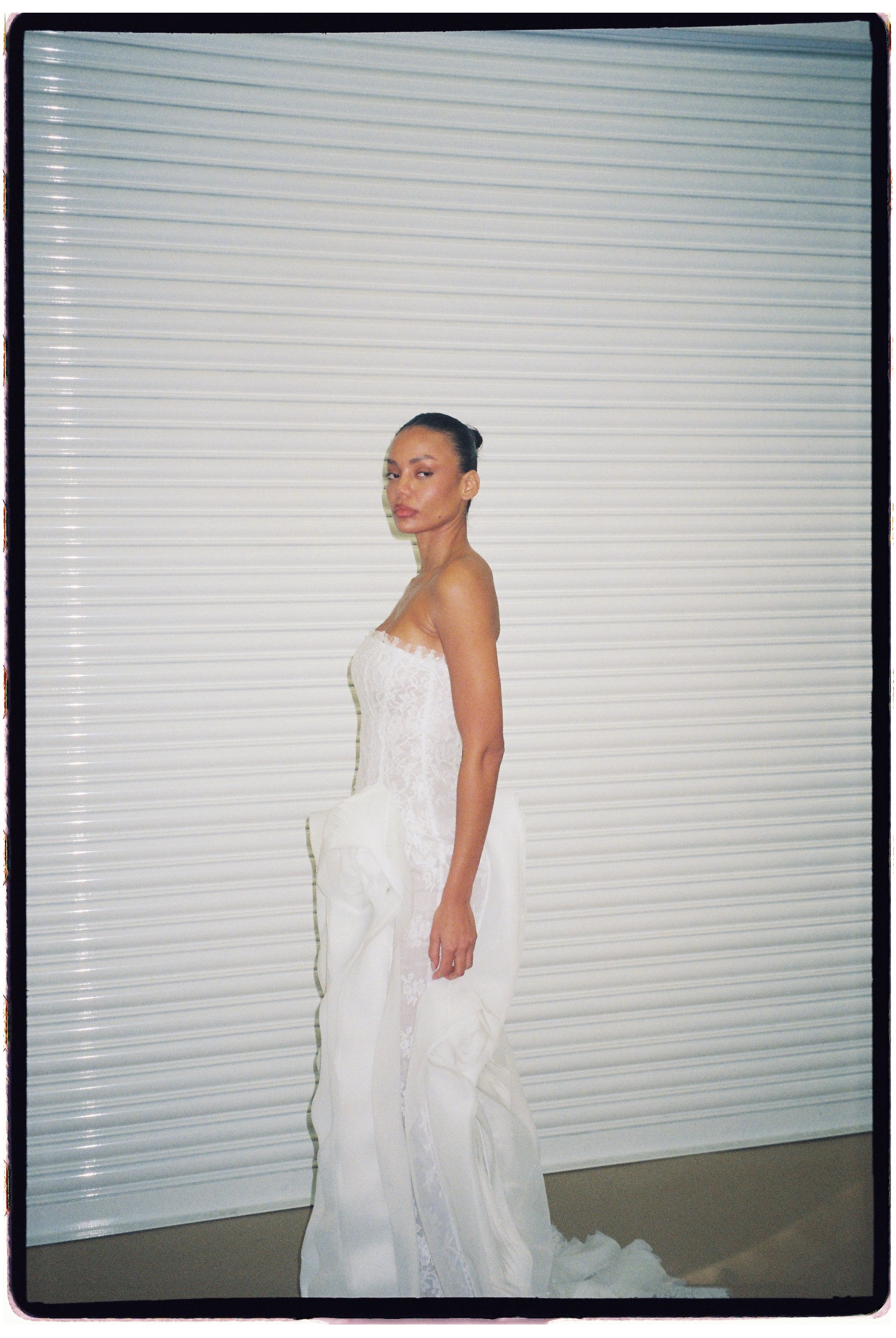 A woman poses in a strapless white bridal gown with ruffled layers against a white slatted backdrop.
