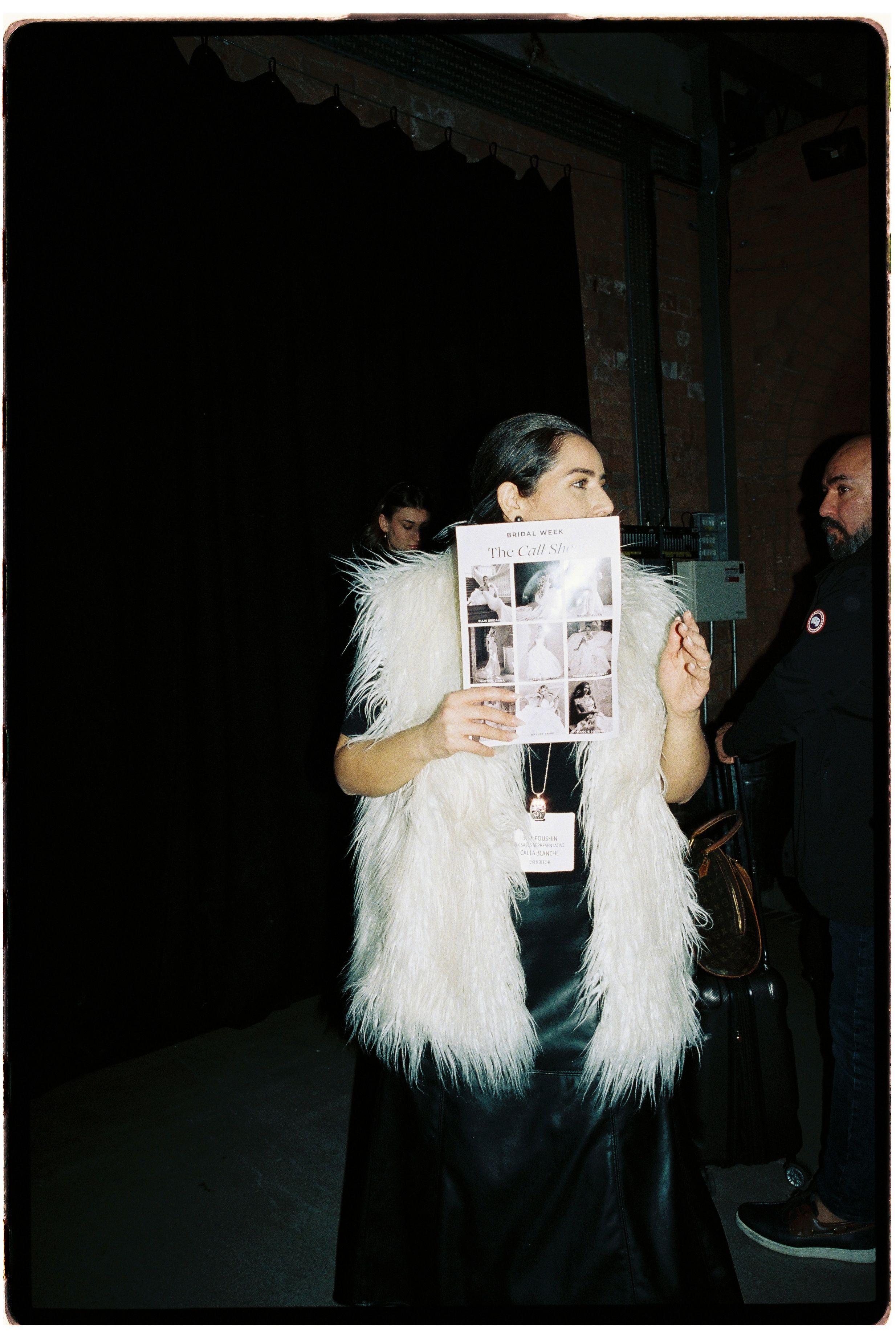 Woman in white faux fur vest holds a magazine at a fashion event, with guests visible in the background.