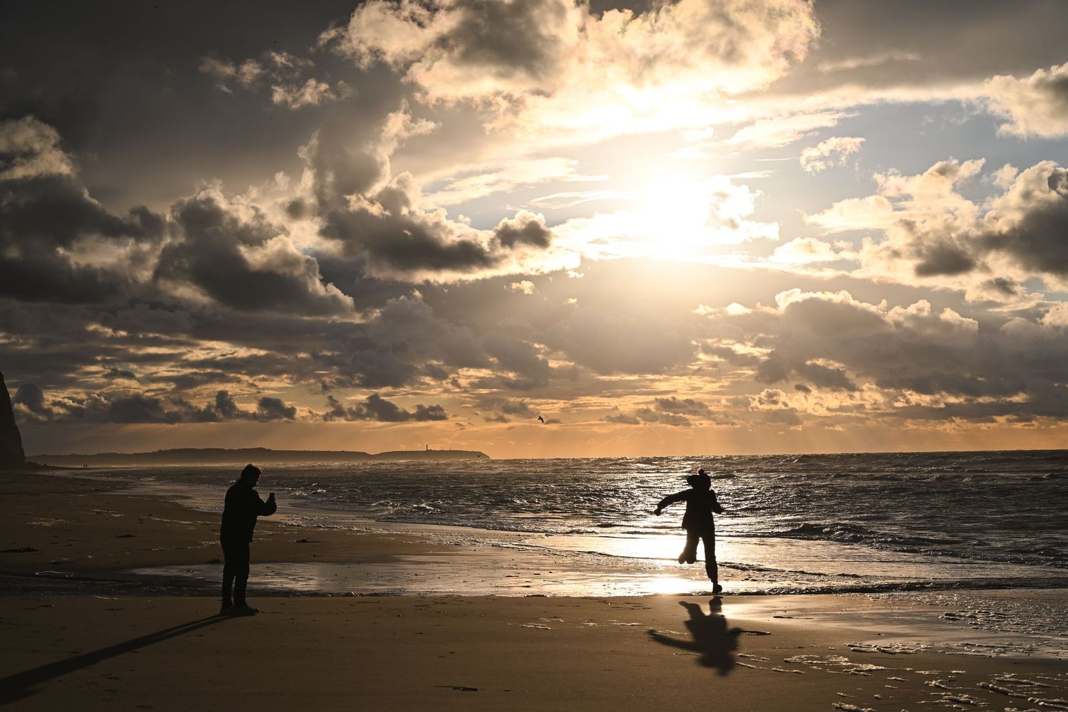 deux silhouettes à contre-jour sur une plage au coucher de soleil, rayons de soleil perçant à travers des nuages spectaculaires