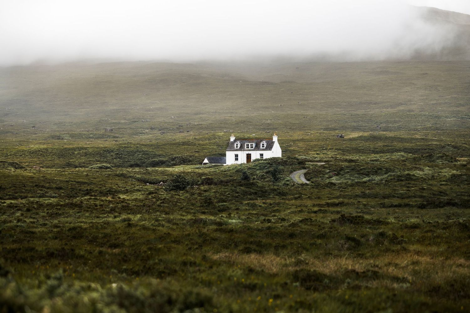 A solitary white cottage sits in a vast misty green highland landscape under a foggy sky.