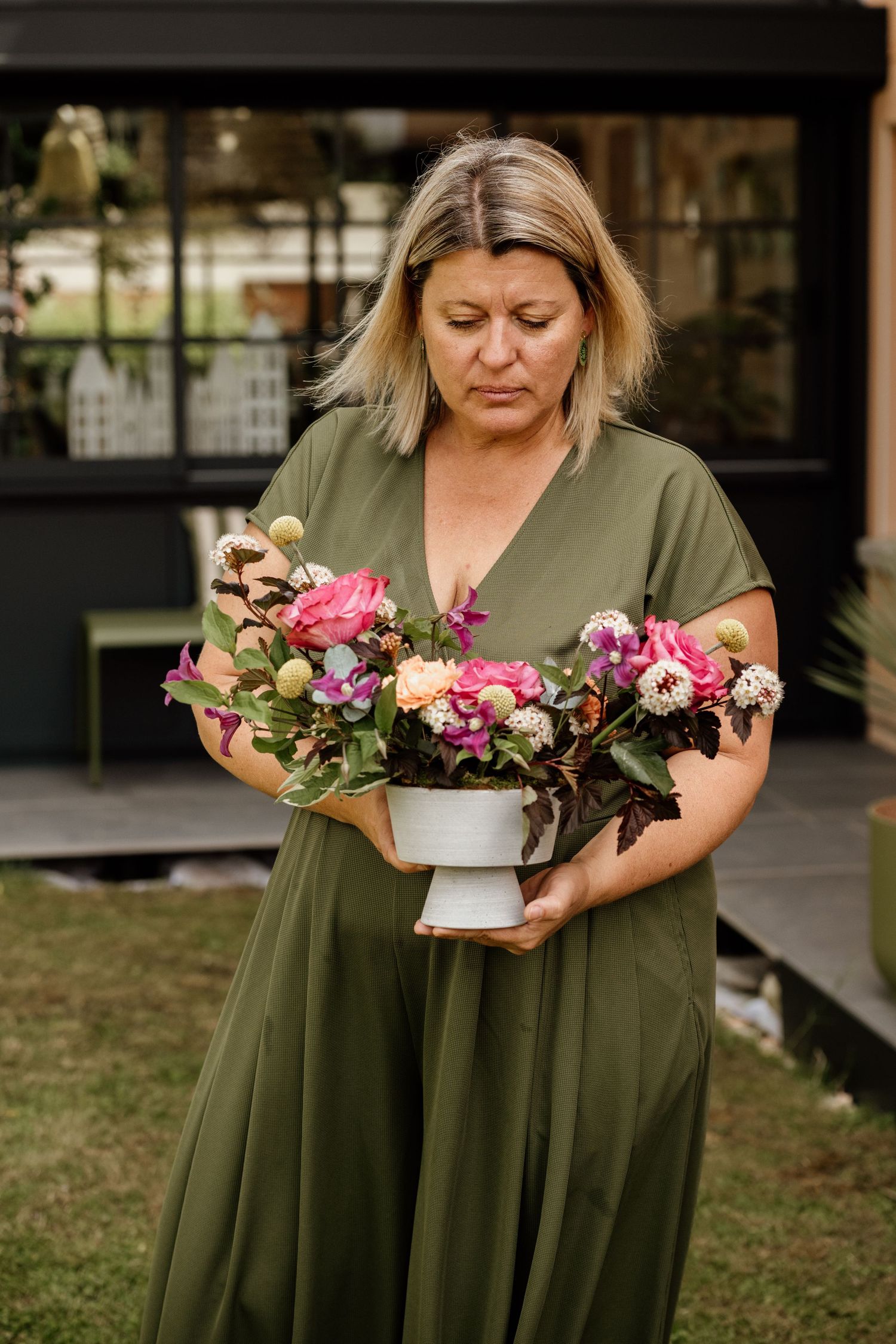 Woman in olive dress holding a floral arrangement with pink peonies and mixed blooms outdoors.