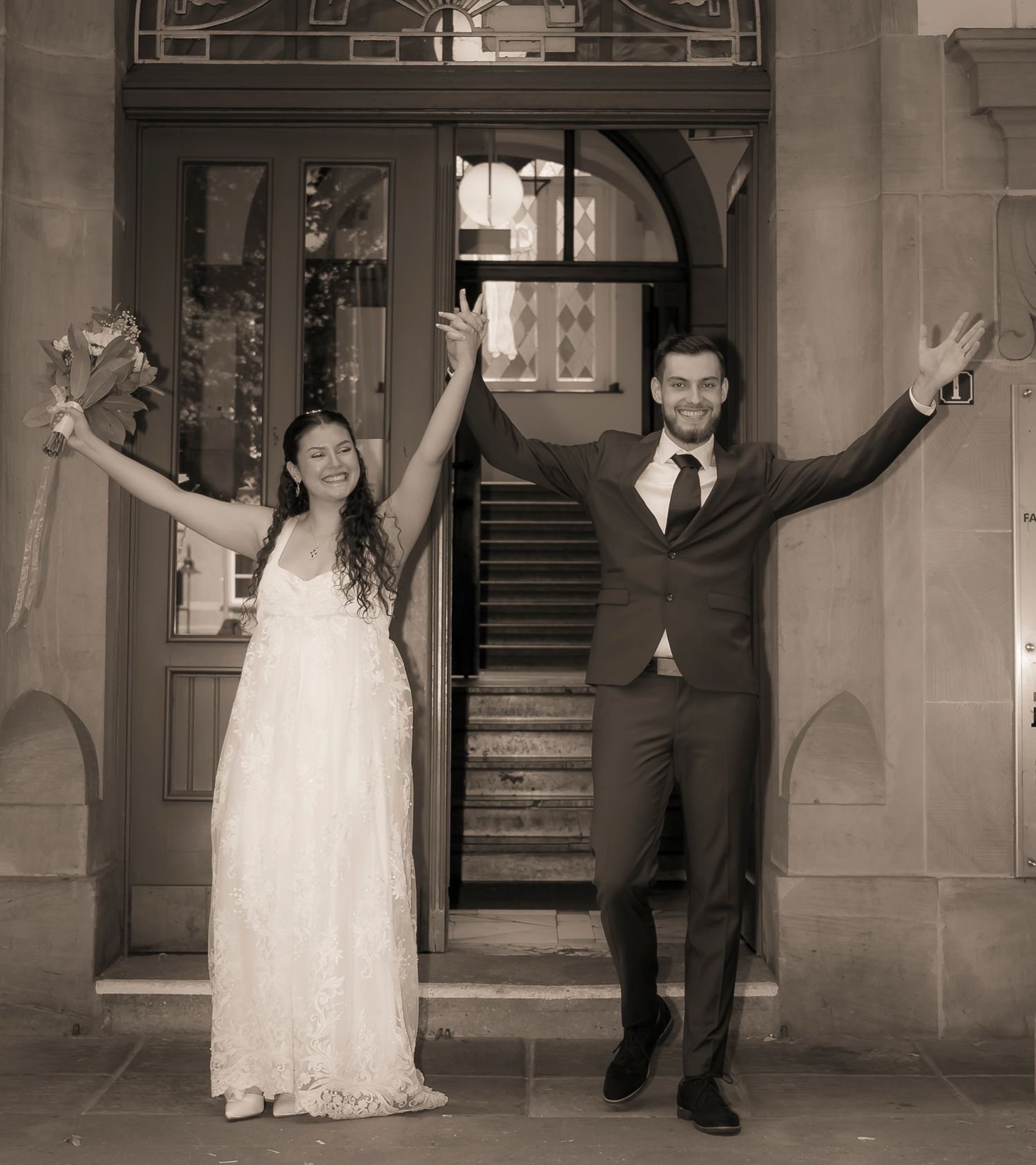Joyful newlywed couple celebrating outside historic stone building in sepia-toned wedding photo.