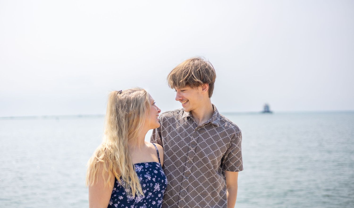 Happy young couple smiling at each other on a bright sunny beach with ocean behind them.