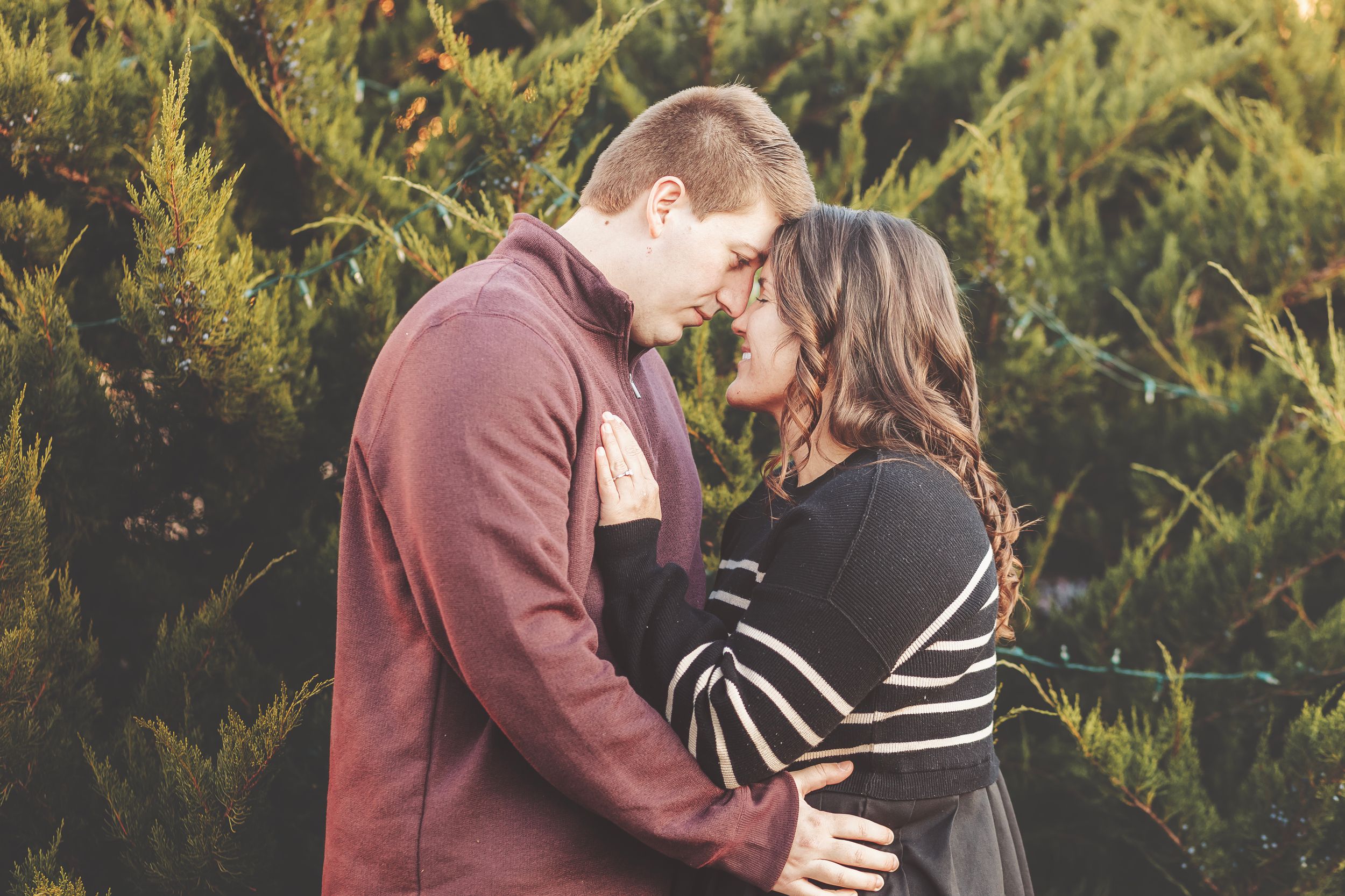 Couple embracing outdoors in golden autumn light, smiling warmly among fall foliage for an engagement photo.