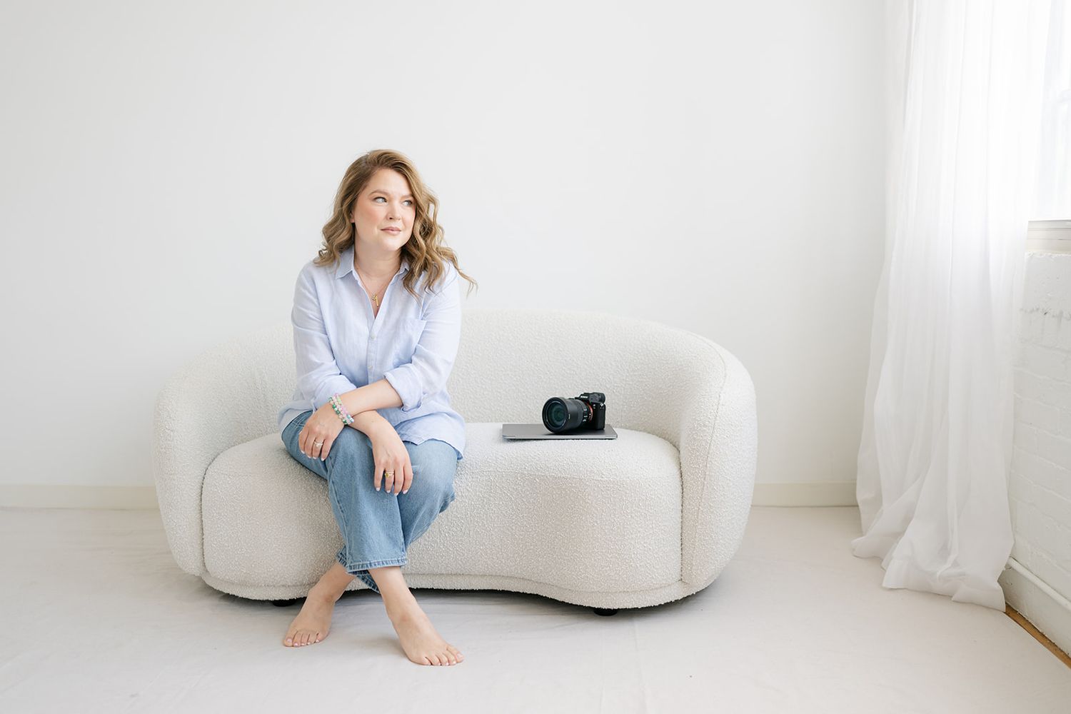 Brand photographer in blue shirt and jeans sits barefoot on white curved sofa with camera beside her in bright minimalist room.
