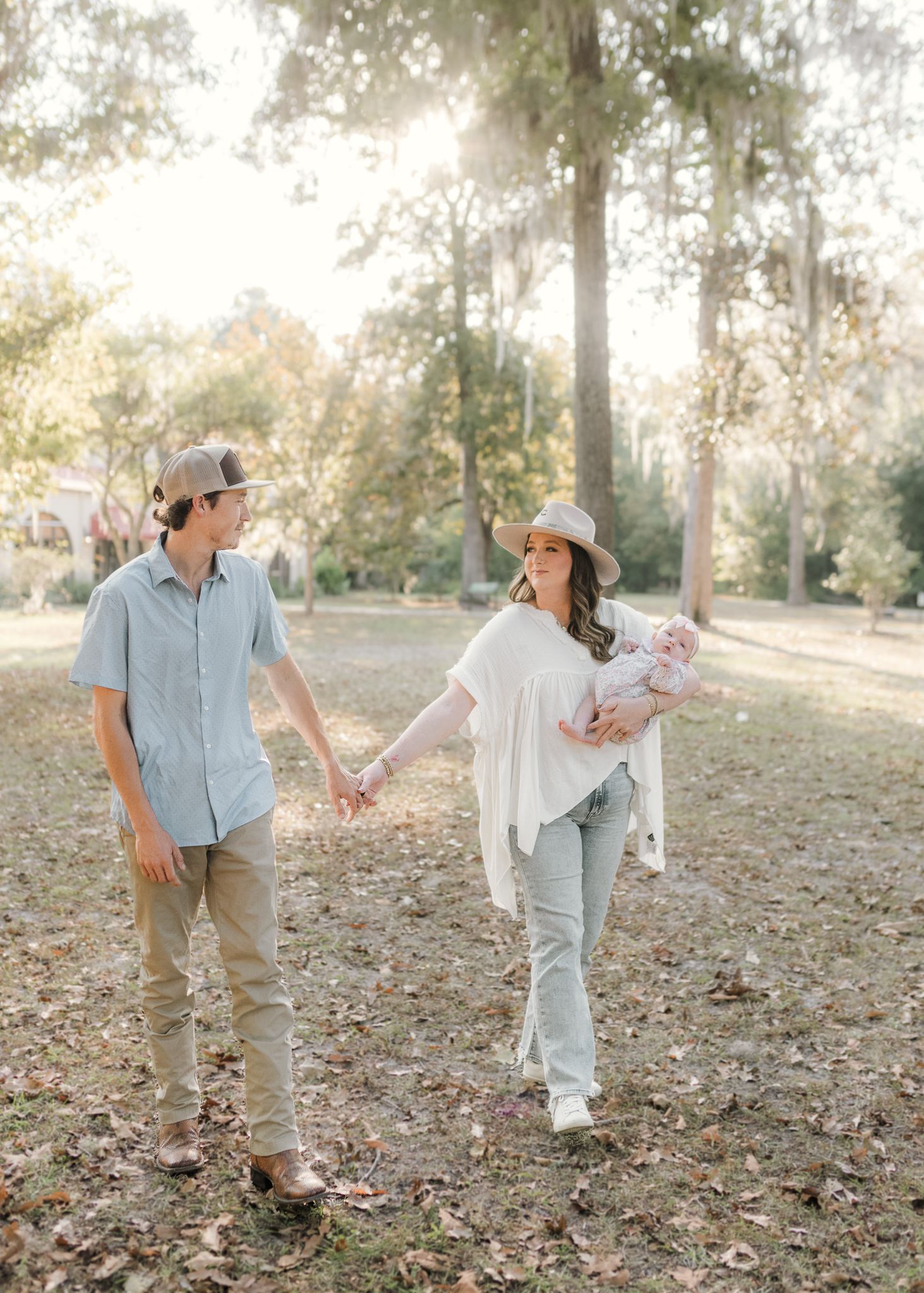 A young couple holding hands walks through a sunlit park, the woman cradling a newborn baby, both wearing casual outfits.