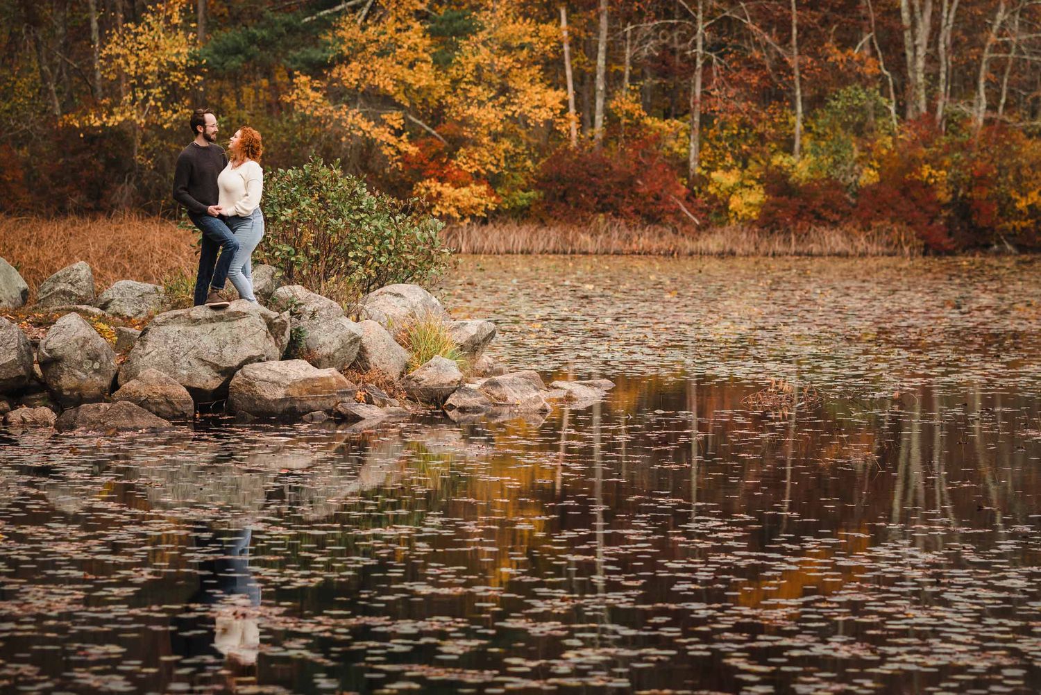 Engaged couple holding hands on a rocky shoreline at Lake Leach in Borderland State Park, surrounded by peak autumn foliage