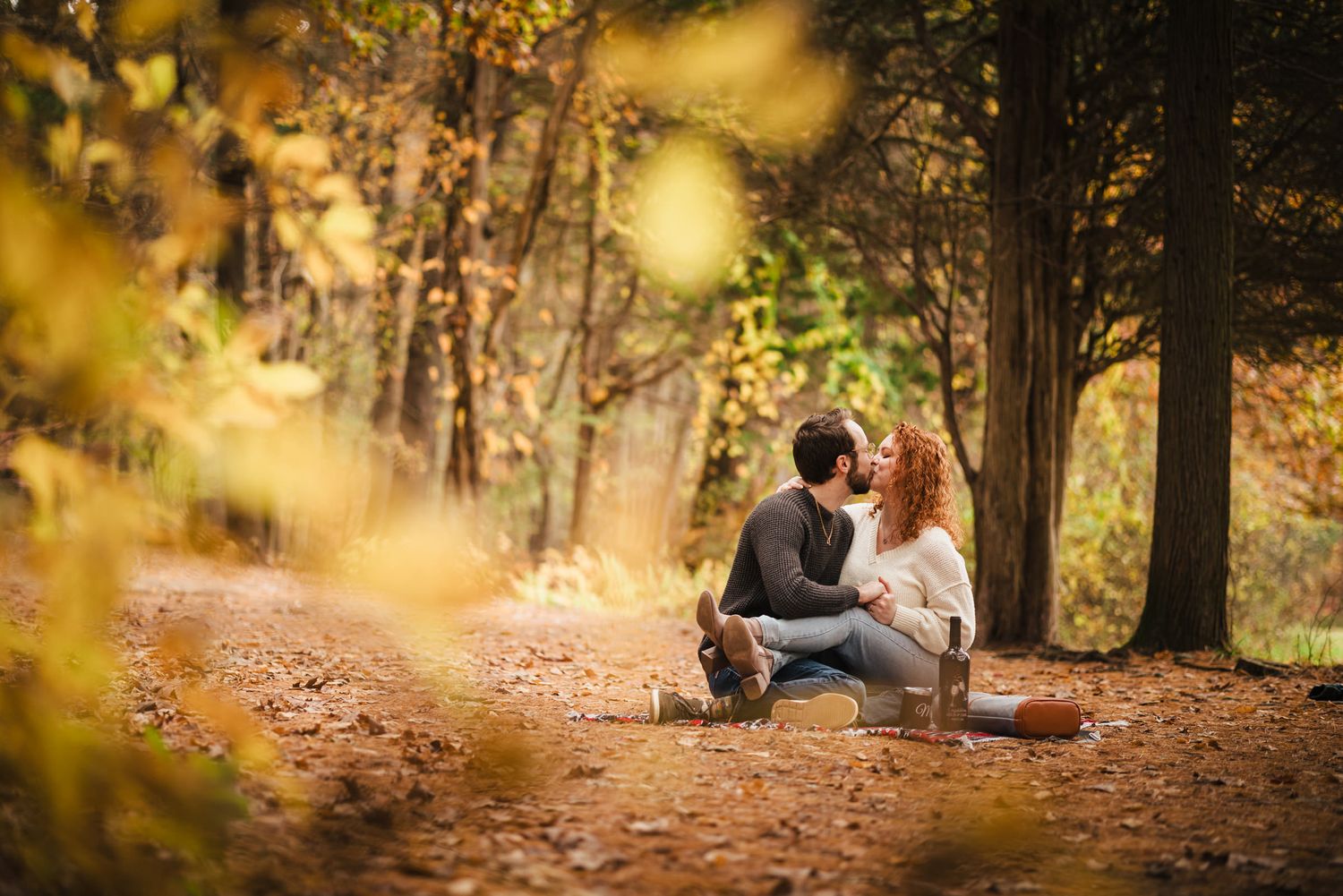 Engaged couple kissing on a picnic blanket in the forest at Borderland State Park, framed by blurred golden leaves in the foreground.