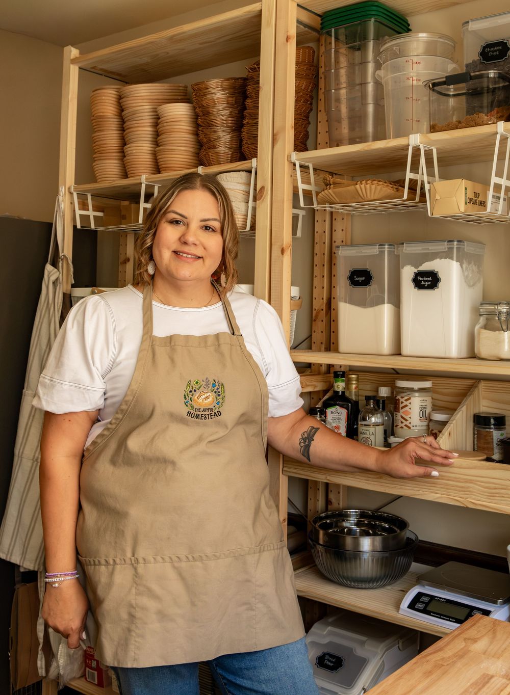 Woman in tan apron stands in organized kitchen pantry with wooden shelves stocked with baking supplies and ingredients.