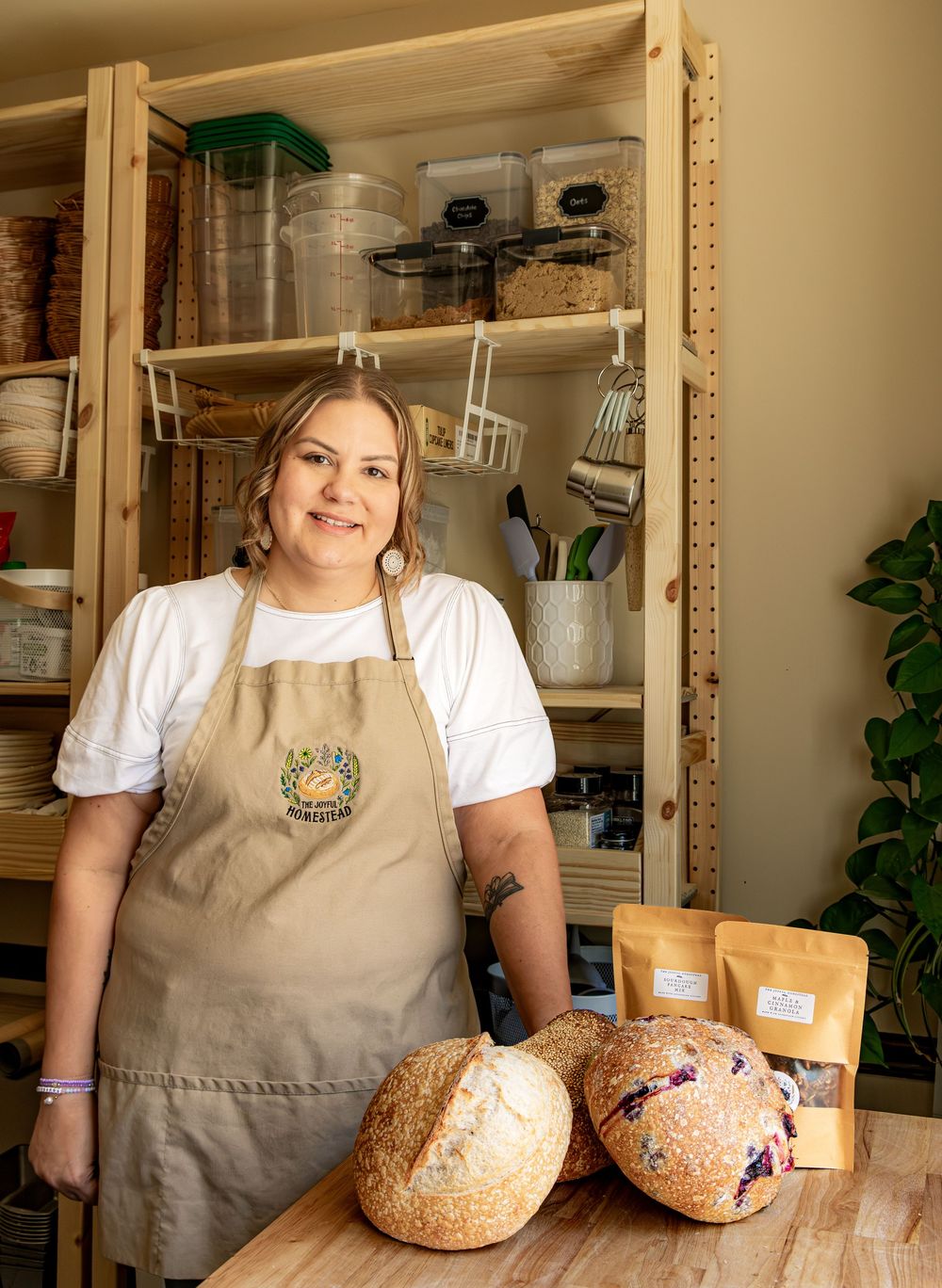 Woman in tan apron stands behind artisan sourdough bread loaves and packaged goods on a wooden table.
