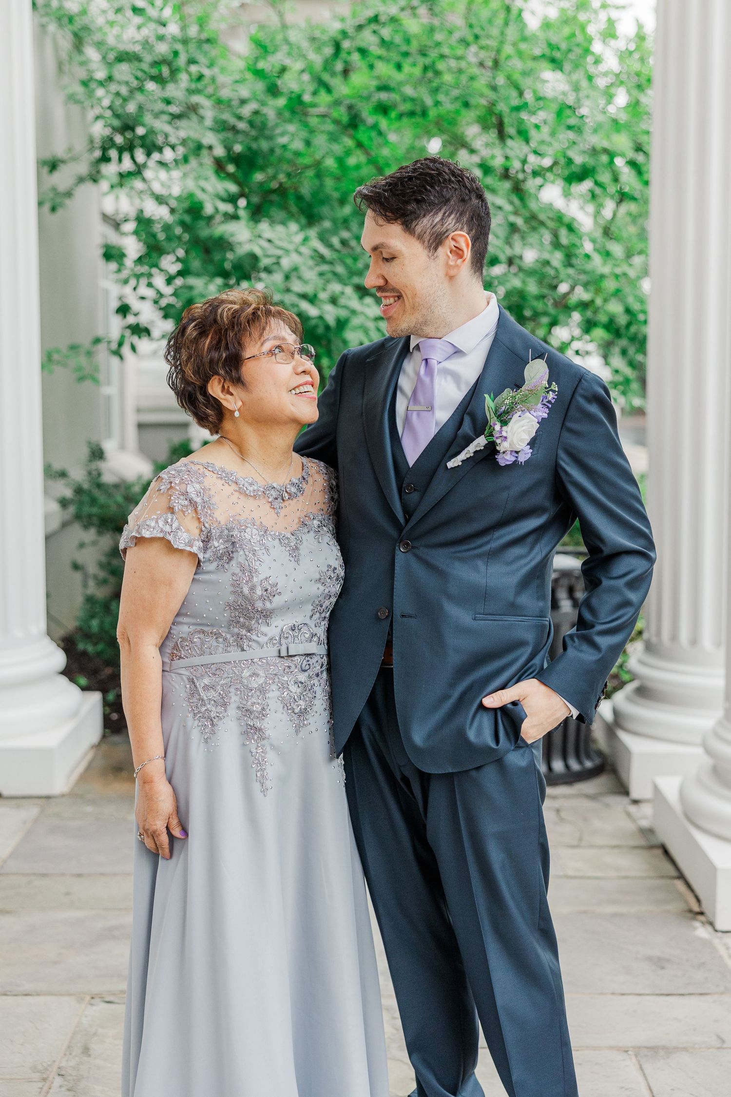 A groom in a navy suit and boutonniere smiles with an older woman in a silver lace gown at a wedding.