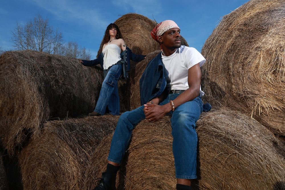 Two models in denim outfits pose on large hay bales outdoors under a blue sky.