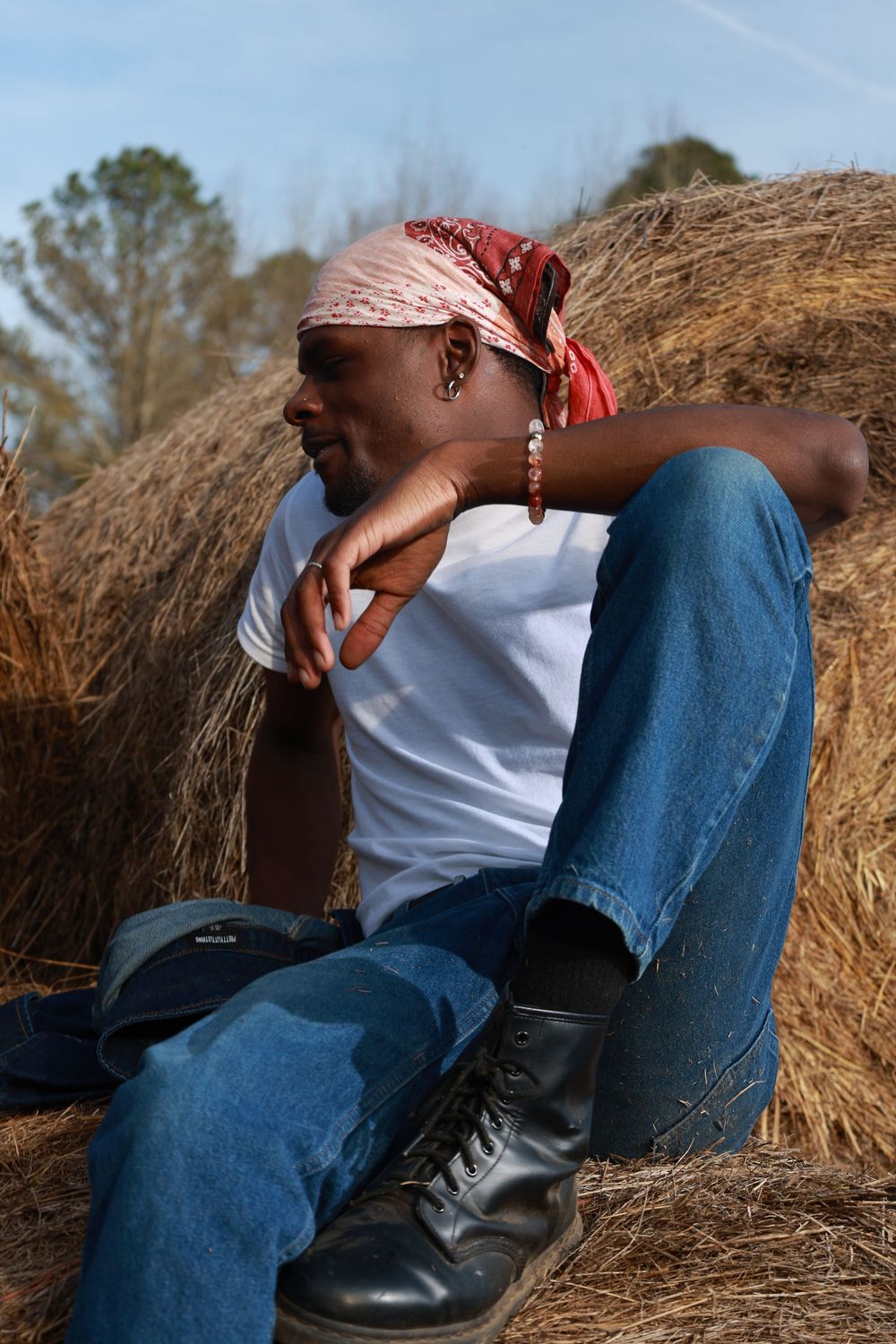 A man in a white t-shirt, jeans, and boots sits against hay bales wearing a red bandana headscarf outdoors.