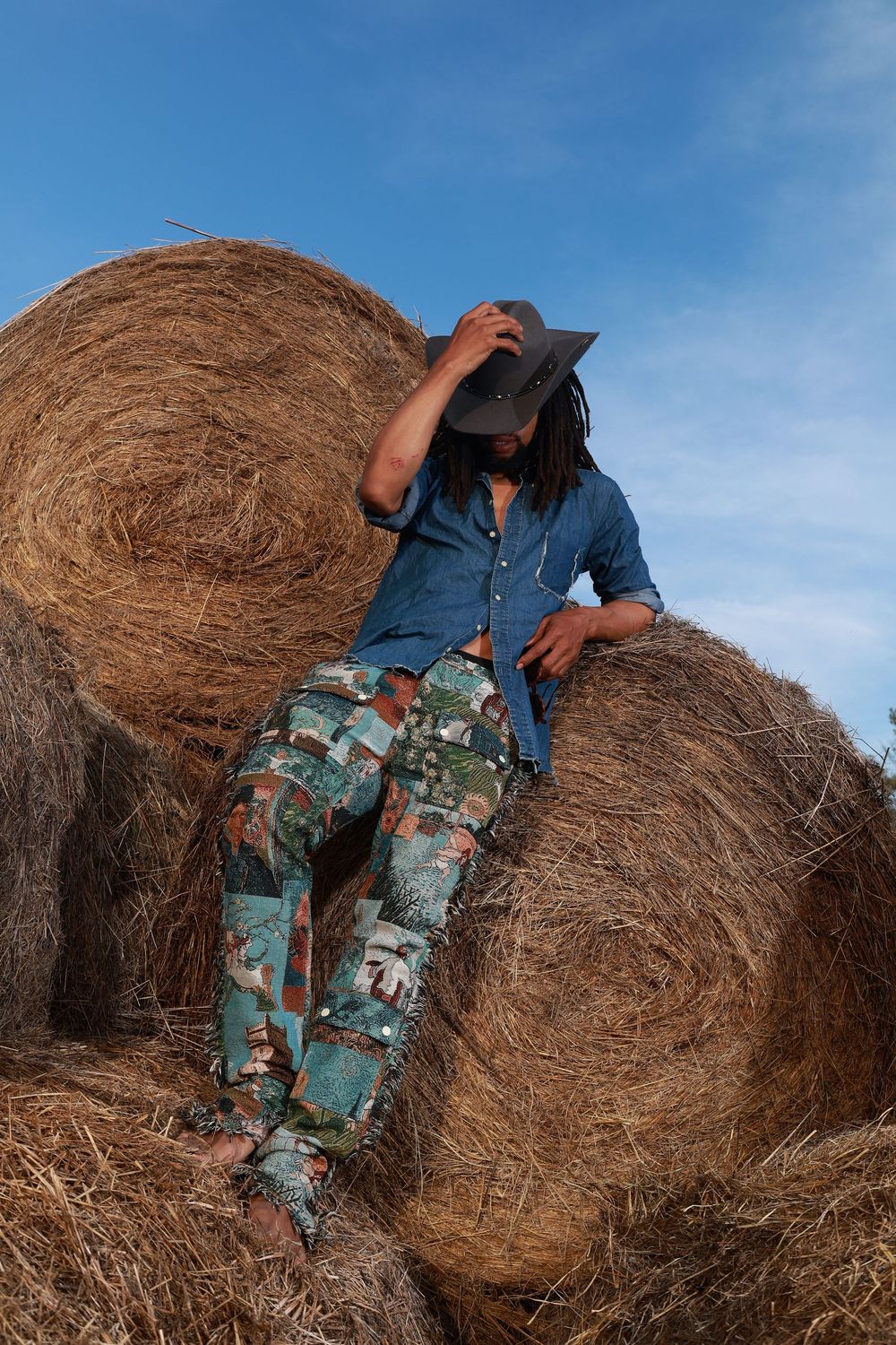 Man with dreadlocks in cowboy hat and camo pants sitting on hay bales against blue sky.