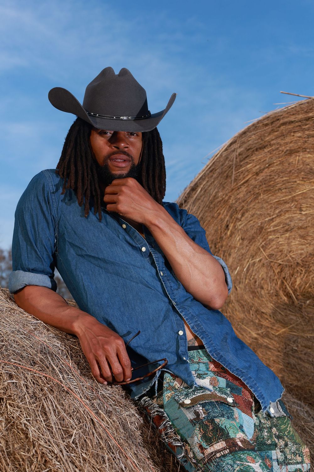 Black man with dreadlocks wearing a cowboy hat and denim shirt leaning against hay bales under a blue sky.