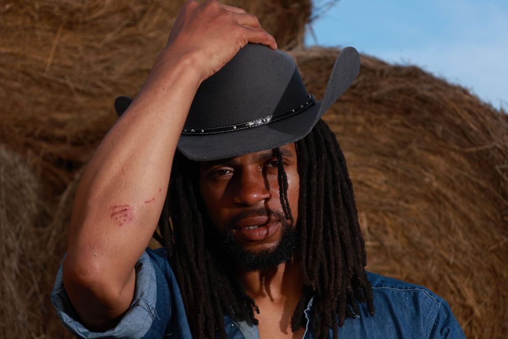 A Black man with dreadlocks wearing a black cowboy hat poses near hay bales in a denim shirt outdoors.