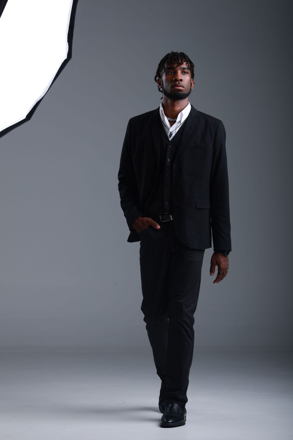 A confident Black man in a black suit walks toward the camera in a professional studio setting.