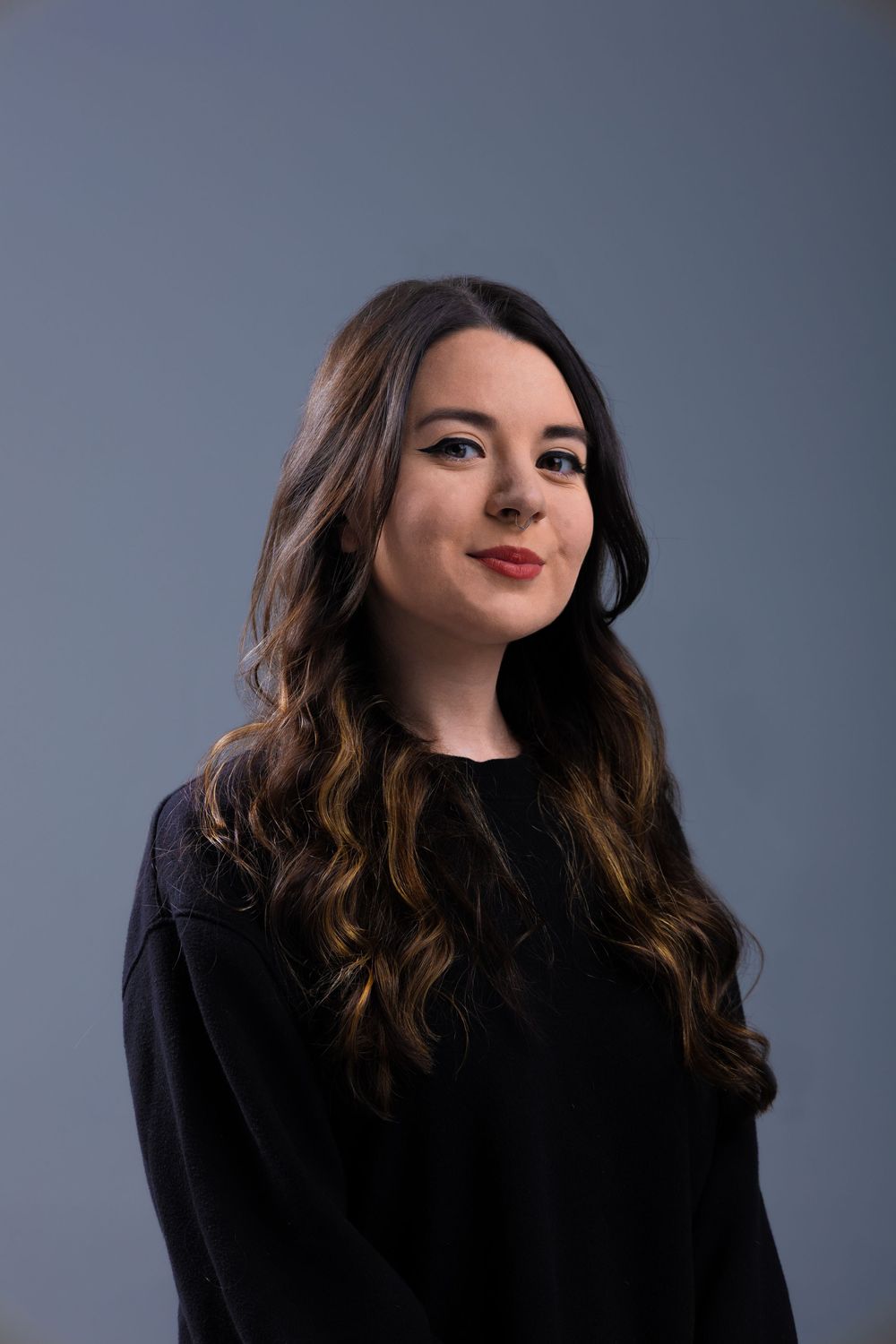 Young woman with long dark wavy hair wearing a black top, posing against a gray studio background.