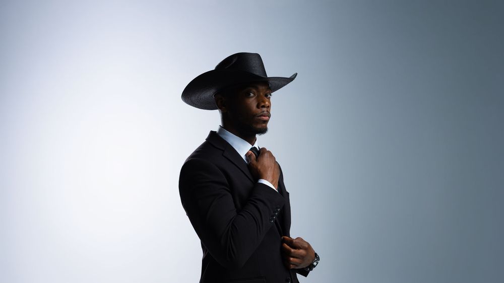 Stylish man in black suit and cowboy hat adjusting tie against gray background.