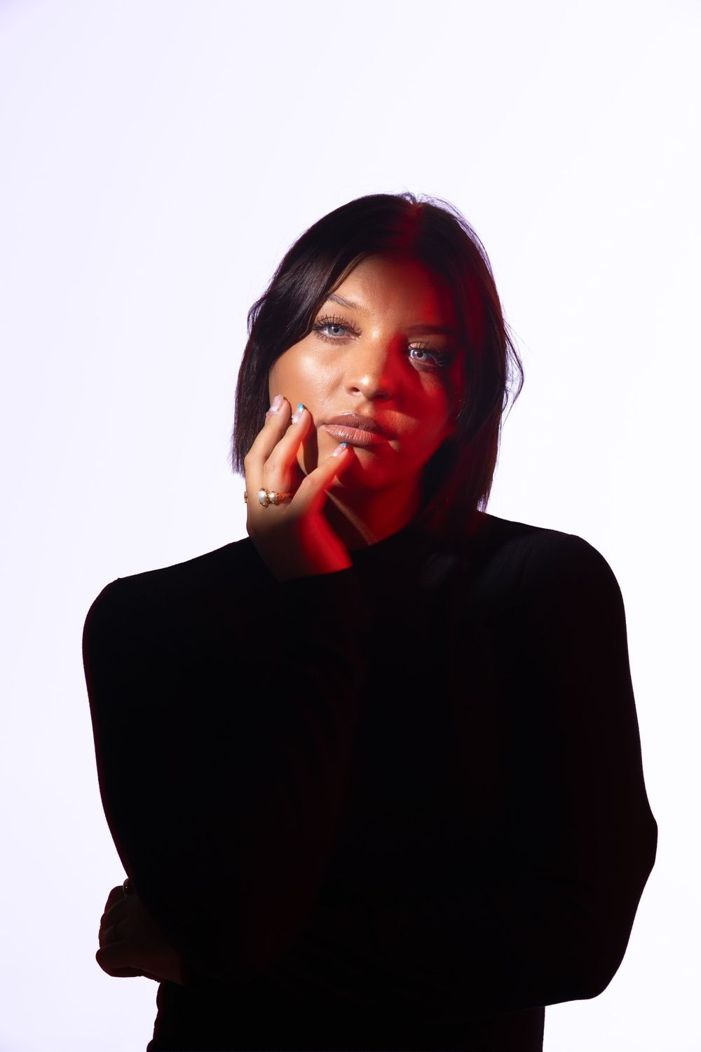 A woman in a black turtleneck poses thoughtfully with red lighting on her face against a white background.