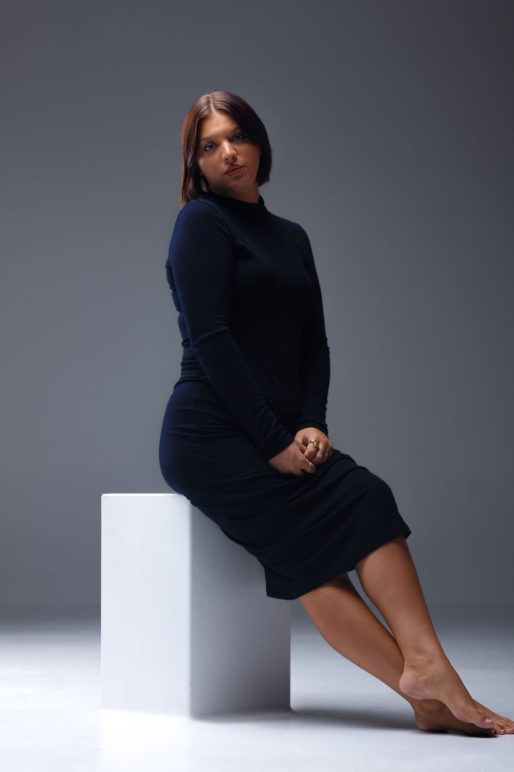 Woman in black long-sleeve midi dress sits barefoot on white cube against gray studio background.