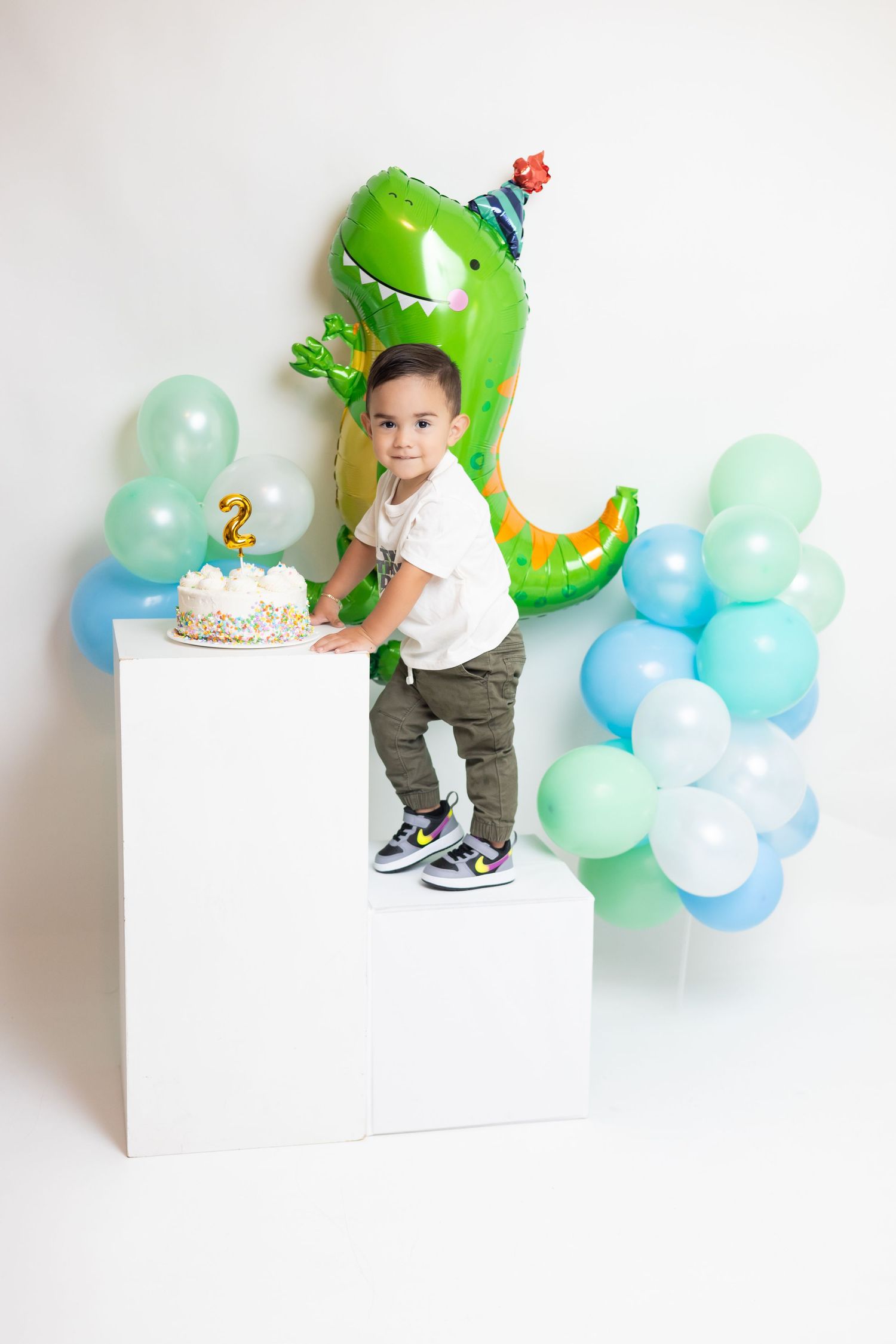 A toddler boy celebrates his 2nd birthday with a dinosaur balloon, cake, and colorful balloons in a white studio setting.