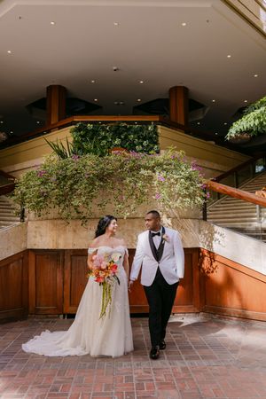 Bride and groom walk hand-in-hand beneath lush floral archway, bride holding vibrant bouquet in elegant off-shoulder gown.