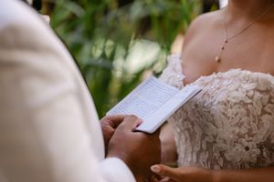 Groom reading wedding vows from a booklet to his bride in a lace dress during an outdoor ceremony.