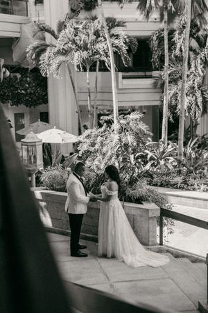 Black and white photo of a bride and groom holding hands in a lush tropical courtyard during their wedding ceremony.