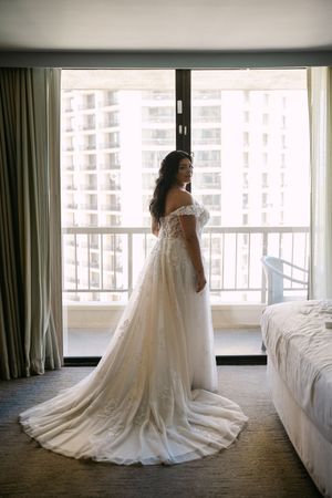 Bride in off-shoulder lace wedding gown with long train stands by hotel window overlooking city skyline.
