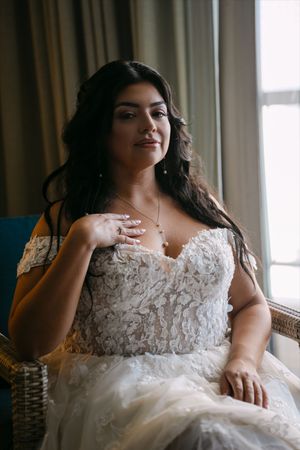 Bride in lace off-shoulder wedding gown sits by window, touching necklace, with elegant updo and soft natural light.