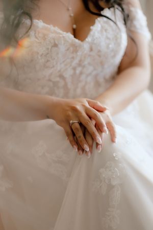 Bride in white lace wedding dress showcasing elegant manicure and diamond engagement ring on her delicate hands.