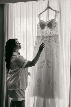 Bride in pajamas admiring her lace wedding dress hanging by a window in a black and white photo.