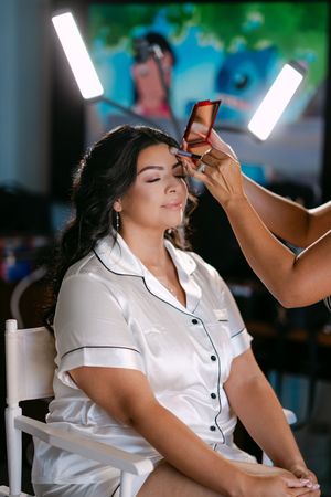 Makeup artist applying eyeshadow to a bride in white satin pajamas under studio lights.