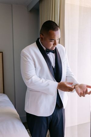 Groom in white tuxedo jacket and black bow tie adjusting cufflinks in a hotel room before wedding ceremony.