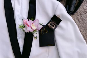 Groom's white tuxedo with pink orchid boutonniere, black suspenders, wedding ring box labeled His, and dress shoes.