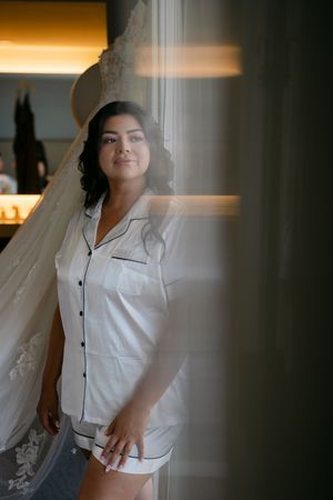 Bride in white satin pajamas standing by a window with her wedding dress hanging in the background.