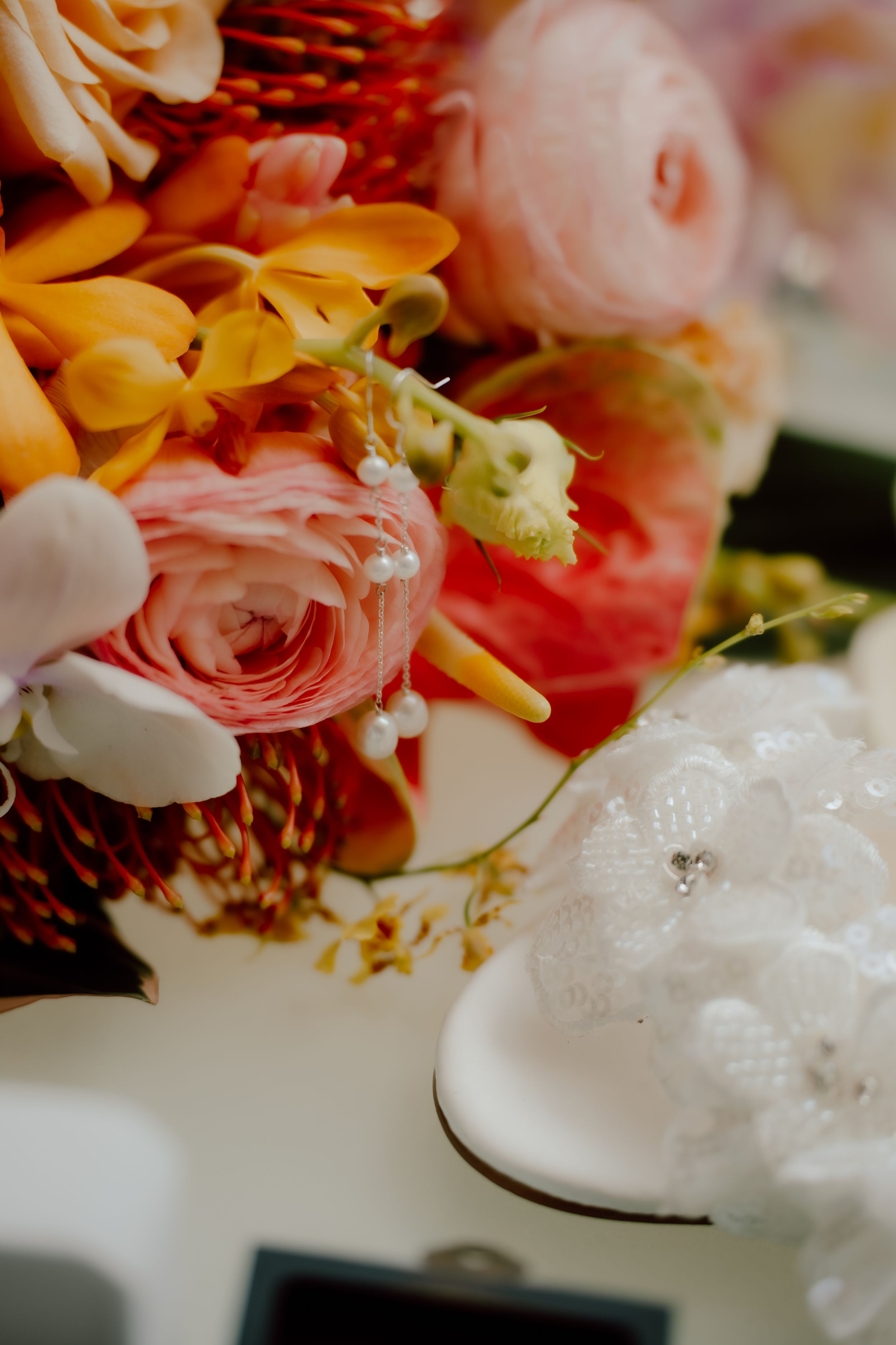 Vibrant bridal bouquet with pink ranunculus, orange orchids, and red berries beside a white lace wedding shoe.