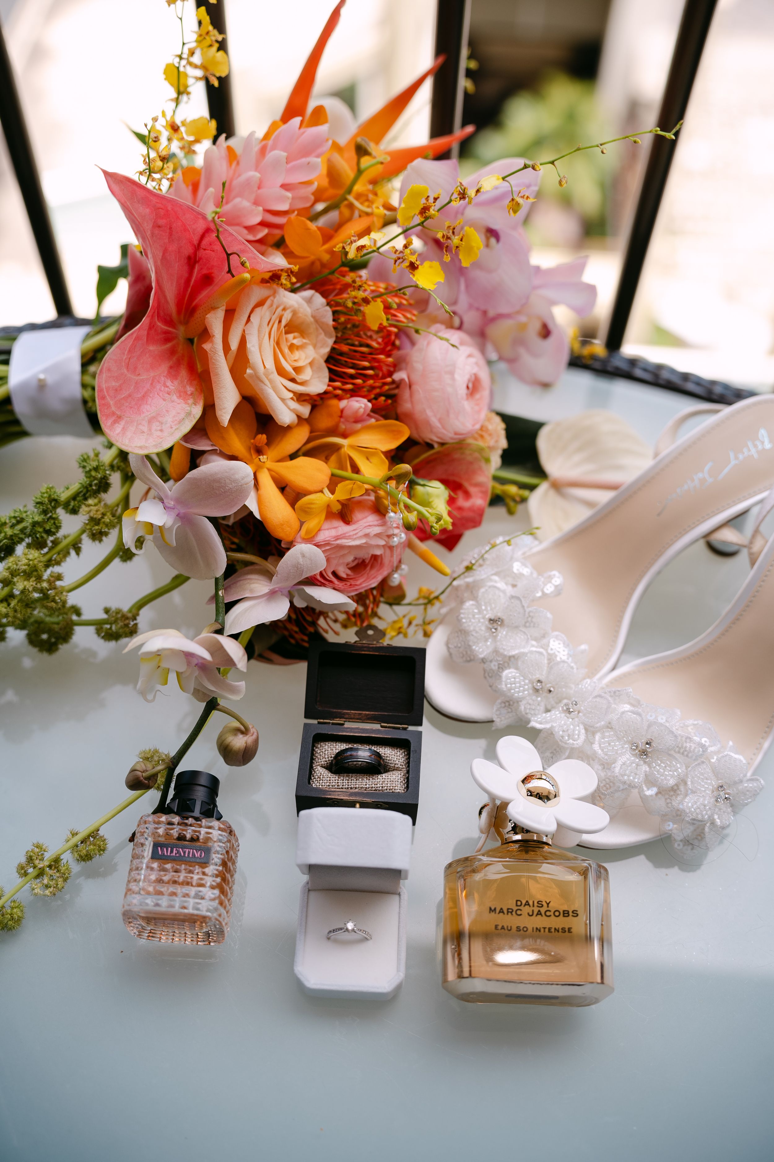 Bridal flat lay with colorful tropical bouquet, perfume bottles, ring box, and white heeled sandals on a light surface.