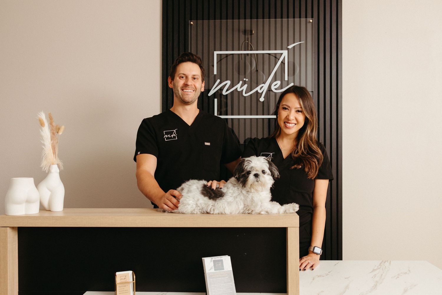 A man and woman in black uniforms smile behind a reception desk at Nude salon, holding a small fluffy dog.