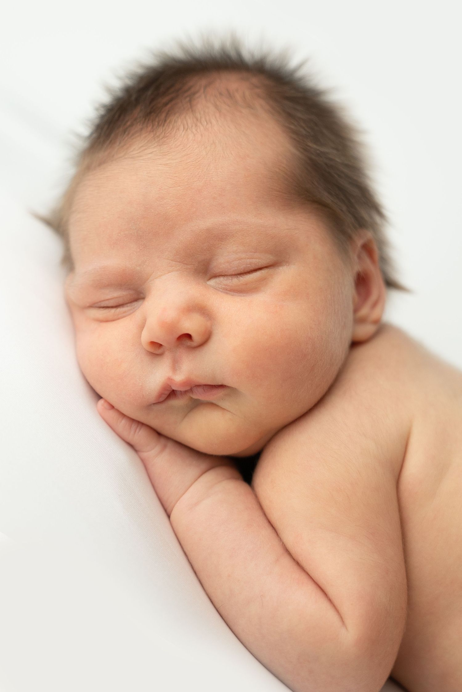 A peaceful newborn baby sleeping with hands tucked under cheek on a soft white background.