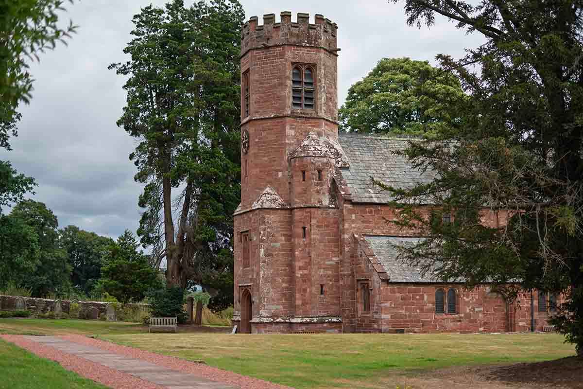 Historic red sandstone church with a round tower surrounded by green trees and manicured lawns under a cloudy sky.